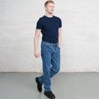 Man wearing a navy t-shirt and blue jeans standing against a white brick wall.