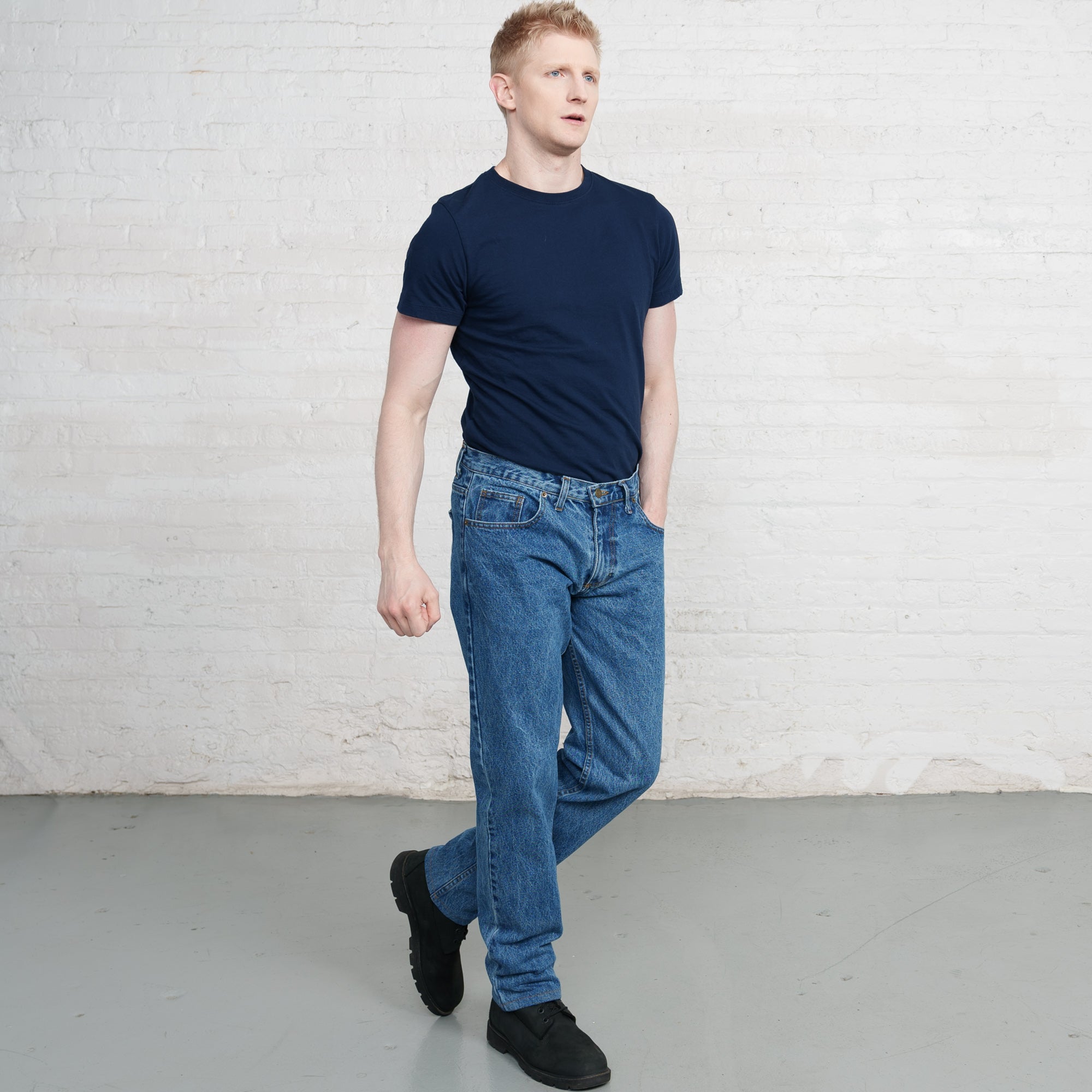 Man wearing a navy t-shirt and blue jeans standing against a white brick wall.